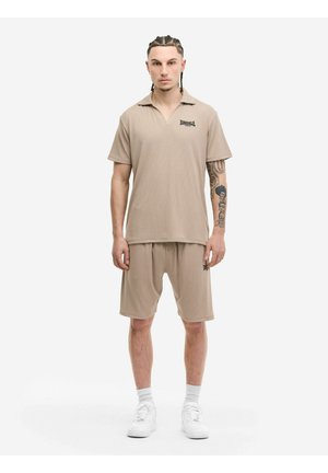 Mann mit geflochtenen Haaren, trägt ein beiges Lonsdale-Poloshirt und passende Shorts, weiße Turnschuhe, steht vor weißem Hintergrund.