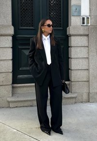 Kobieta w czarnym garniturze, białej koszuli, białym krawacie, czarnych okularach przeciwsłonecznych i złotych kolczykach stoi przed ciemnozielonymi drzwiami, trzymając czarną torebkę.