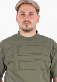 Zielona bawełniana koszulka z podniesionym prostokątnym wzorem oraz wyraźnym poziomym paskiem. Krótkie rękawy i okrągły dekolt.