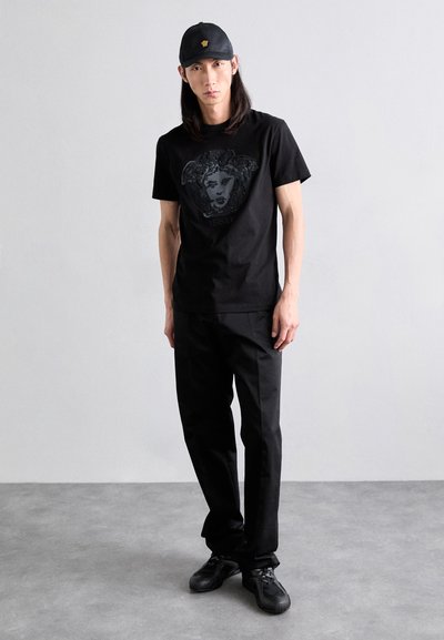 Black T-shirt with a textured graphic design, paired with black trousers and black shoes. The hat is also black, featuring a logo.