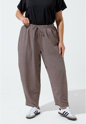 Persona che indossa pantaloni larghi a righe con coulisse in vita, t-shirt nera e scarpe da ginnastica bianche con righe nere, in piedi su sfondo uniforme.