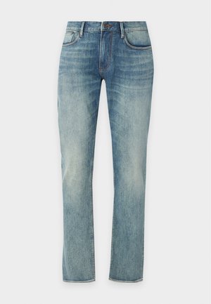 Jeune homme portant une chemise blanche à manches longues avec un imprimé graphique noir, jean bleu, chaussures noires, debout avec une main dans la poche sur fond uni.
