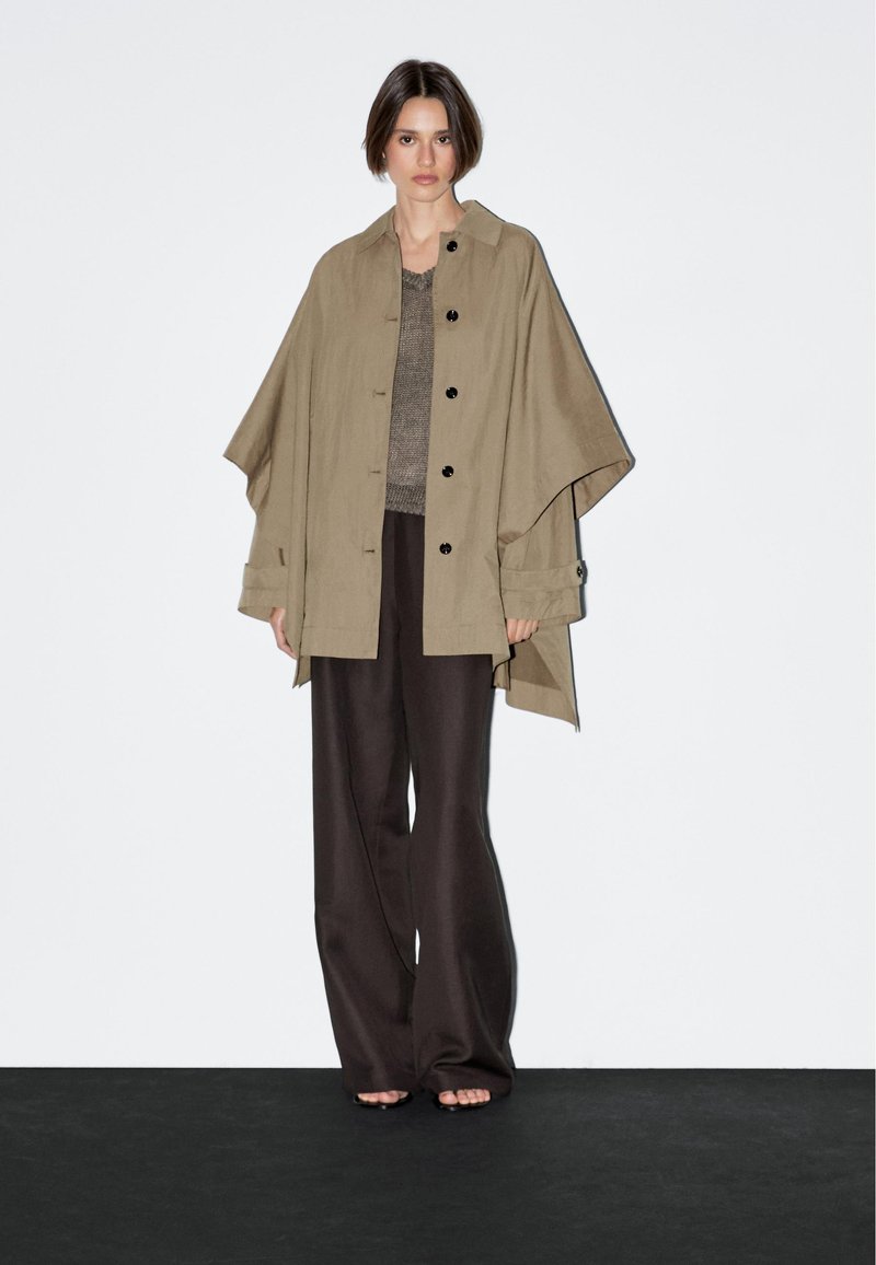 Beige Übergröße Button-Up-Oberbekleidung mit mehrfarbigen Knöpfen, getragen über einem strukturierten grauen Oberteil und dunkelbraunen weiten Hosen.