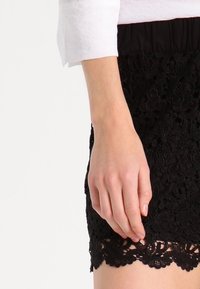 Czarna koronkowa spódnica z strukturalnym kwiatowym wzorem, z elastycznym pasem. Ręka znajduje się blisko dołu, ukazując szczegóły koronkowego wykończenia.