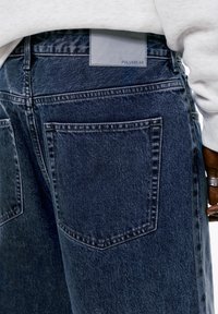 Jeans in denim blu scuro con una texture liscia, caratterizzati da due tasche posteriori e un'etichetta in pelle con il marchio "PULL&BEAR" sulla vita.