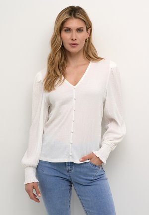Weiße, strukturierte Bluse mit V-Ausschnitt, Knopfleiste und langen, gepufften Ärmeln. Die Bündchen haben ein gerafftes Design. Getragen mit Jeans.