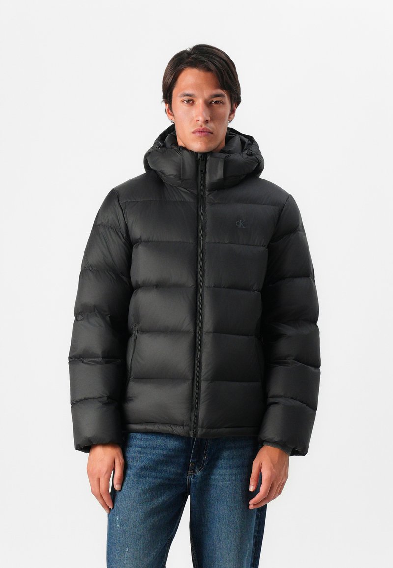 Zwarte pufferjack met een capuchon, voorzien van gestikte secties en een rits aan de voorkant. Het materiaal lijkt glad met een matte afwerking.