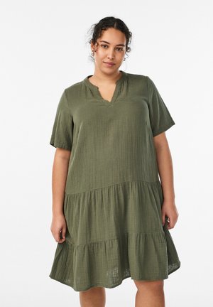 Kobieta z kręconymi włosami, ubrana w luźną, oliwkowozieloną sukienkę na krótki rękaw z warstwami, stojąca na tle gładkiego jasnego tła.