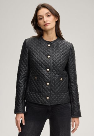 Frau trägt eine schwarze gesteppte Jacke mit goldenen Knöpfen und Vordertaschen, kombiniert mit schwarzen Jeans, vor einem schlichten hellen Hintergrund.