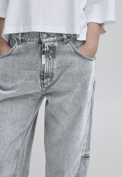 Gray denim jeans feature a relaxed fit, distressed texture, and side pockets. The button closure and brand label are visible at the waist.