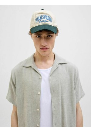 Kortærmet, lysegrøn tekstureret skjorte med knapper, båret over en hvid T-shirt, parret med en beige og grøn kasket med tekst.