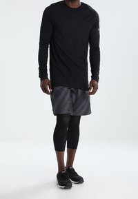 Pitkähihainen musta urheilupaita, jossa on huomaamaton raidallinen muotoilu, yhdistettynä harmaan raidallisiin shortseihin ja mustiin kompressiolaihin. Mustat lenkkarit viimeistelevät asun.