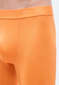 Oranžové priliehavé nohavičky s textúrovaným vzorom a pásom s logom značky. Tkanina sa zdá byť ľahká a pružná.