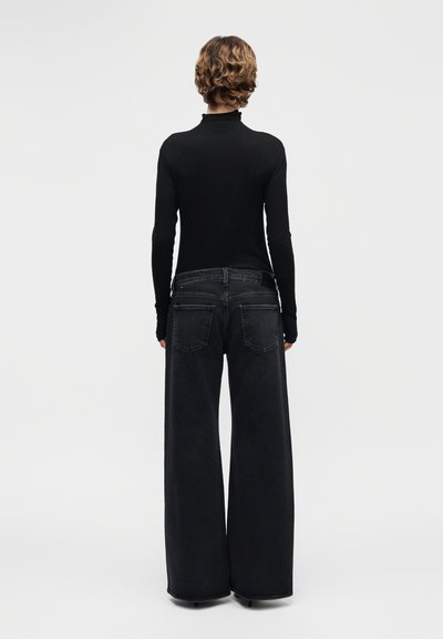 Άτομο με κοντά σγουρά μαλλιά, που φοράει μαύρο ζιβάγκο και φαρδιά μαύρα τζιν παντελόνια, στέκεται γυρισμένο προς τα πίσω μπροστά σε απλό φόντο.