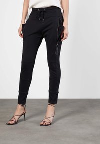 Schwarze Jogginghosen aus weichem Material, mit Reißverschlusstaschen, tapered Design und Bündchen am Knöchel. Kombiniert mit schwarzen, schmalen High Heels.