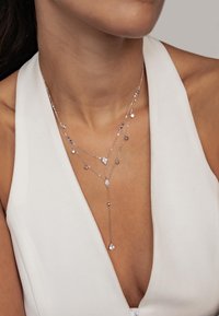 Srebrny naszyjnik z warstwowym wisiorem, składający się z kilku łańcuszków, ozdobiony akcentami w kształcie kropli i okręgów, spoczywający na nagiej szyi.