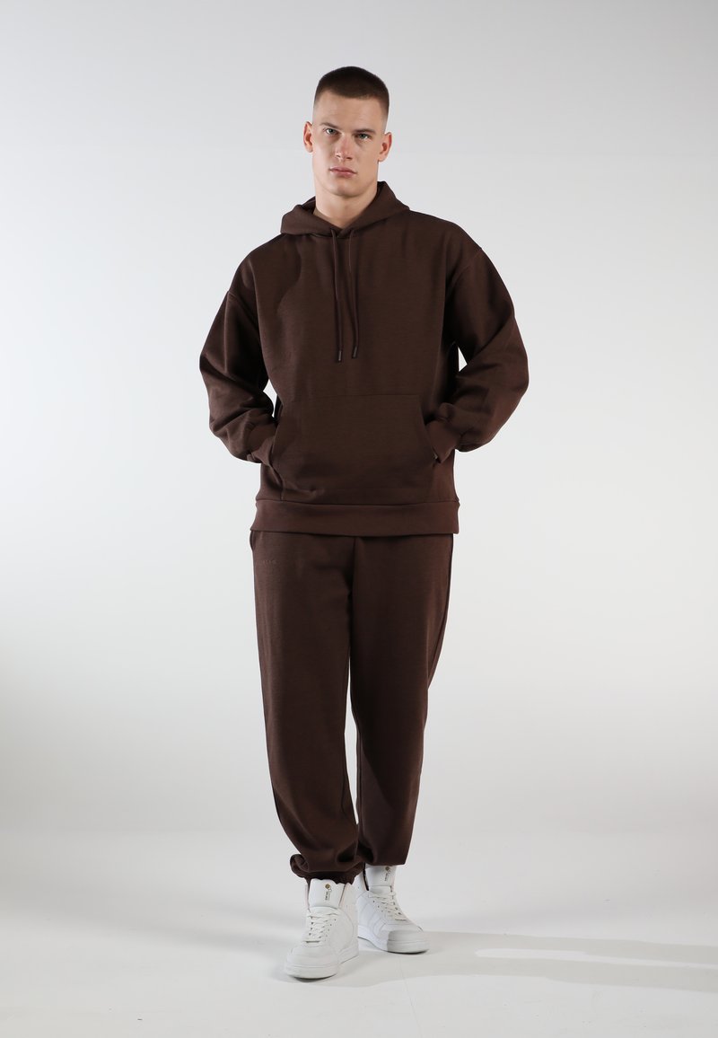 Brauner Hoodie und Jogginghose aus weichem Stoff mit lockerer Passform. Verfügt über eine Kängurutasche und eine Kapuze mit Kordelzug. Kombiniert mit weißen Sneakern.