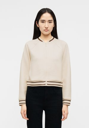 Mladá žena s dlhými tmavými vlasmi, oblečená v béžovej bunde na zips s hnedými pruhovanými manžetami a lemom, stojaca na jednoduchom svetlom pozadí.