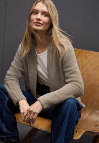Cardigan beige, texturé, avec de larges revers, porté sur une chemise blanche et un jean bleu marine à jambe large, assis sur une chaise en bois devant un fond gris.