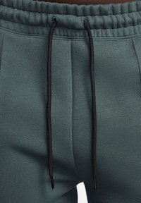 Donkergroene sweatpants van een textuurstof, met een elastische tailleband en een zwarte trekkoord. Heeft zijzakken en subtiele naaddetails.