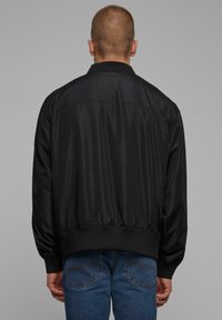 Veste bomber noire avec une texture lisse et brillante, présentant un ourlet et des poignets côtelés. Pas de marquage visible. Vue de dos, avec un jean bleu décontracté.