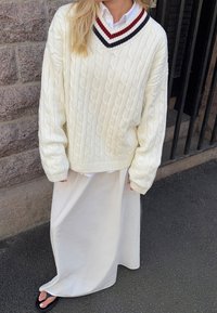 Cremfarbener, grobstrickiger Pullover mit V-Ausschnitt, akzentuiert mit kastanienbraunen und marineblauen Streifen; kombiniert mit einem fließenden weißen Rock und schwarzen Sandalen.
