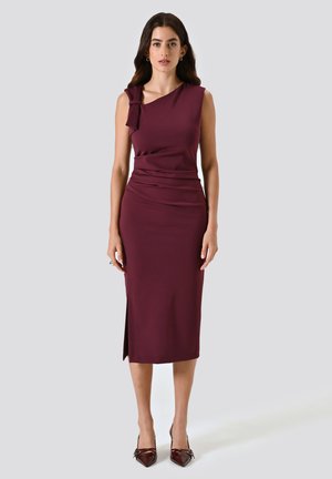Robe midi bordeaux avec un design asymétrique à une épaule, décorée d'un nœud. Tissu lisse, fente sur le côté et silhouette ajustée. Talons aiguilles visibles.