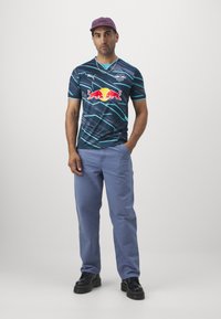 Sötétkék sportmez, teal csíkokkal, a középpontjában egy hangsúlyos Red Bull logóval. Világoskék nadrággal és fekete cipővel párosítva.