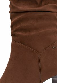 Stivale alla caviglia in suede marrone con parte superiore arricciata, dettagli di cucitura discreti, punta affusolata e tacco sottile. Accento di hardware minimalista in argento.