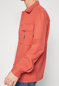 Man i röd långärmad skjorta med knappar och bröstfickor samt blå jeans, sedd från sidan mot en neutral bakgrund.