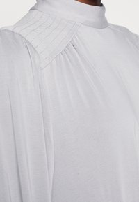 Blouse gris clair avec un col haut, des détails plissés aux épaules et un tissu lisse et fluide. Présente une finition douce et texturée.