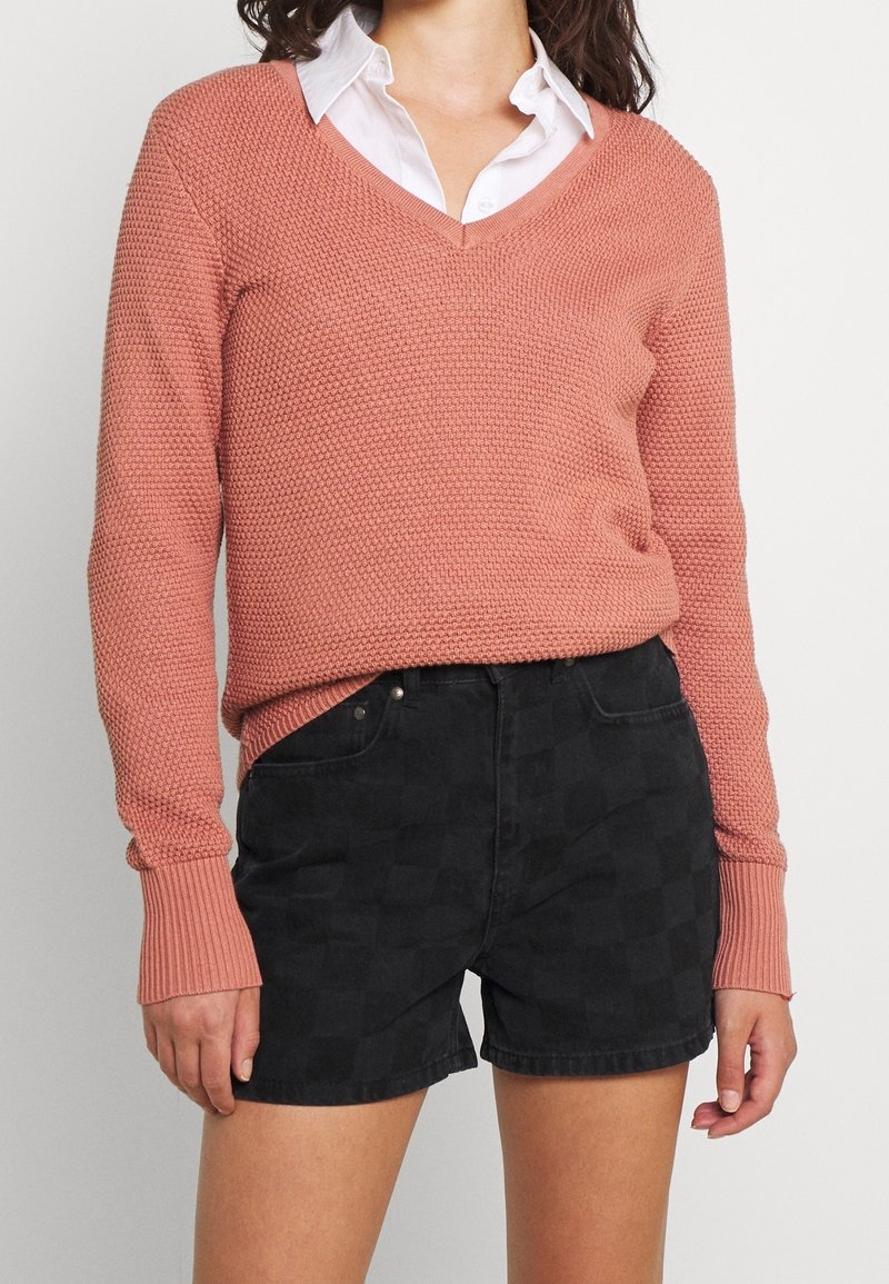 Hellrosa strukturierter Strickpullover mit V-Ausschnitt und langen Ärmeln, kombiniert mit schwarzen karrierten Jeans-Shorts mit Vorderentaschen.
