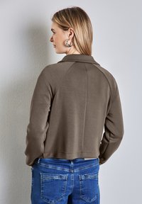 Brun kort jacka med krage, med en slät textur och raglanärmar, parat med blåjeans som har detaljerad sömnad.