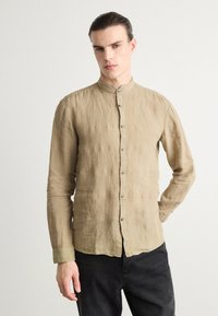 Camisa de lino beige con cuello alto, mangas largas y botones plateados. Tejido texturizado con un corte relajado y dobladillo curvado.