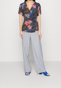 Top blu con motivo floreale e maniche corte, abbinato a pantaloni a gamba larga grigio chiaro e sandali con tacco aperti blu.