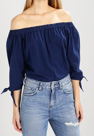 Kobieta ubrana w granatową bluzkę z odkrytymi ramionami i rękawami do łokcia, związanymi przy mankietach, w połączeniu z jasnoniebieskimi, postrzępionymi jeansami z wysokim stanem.