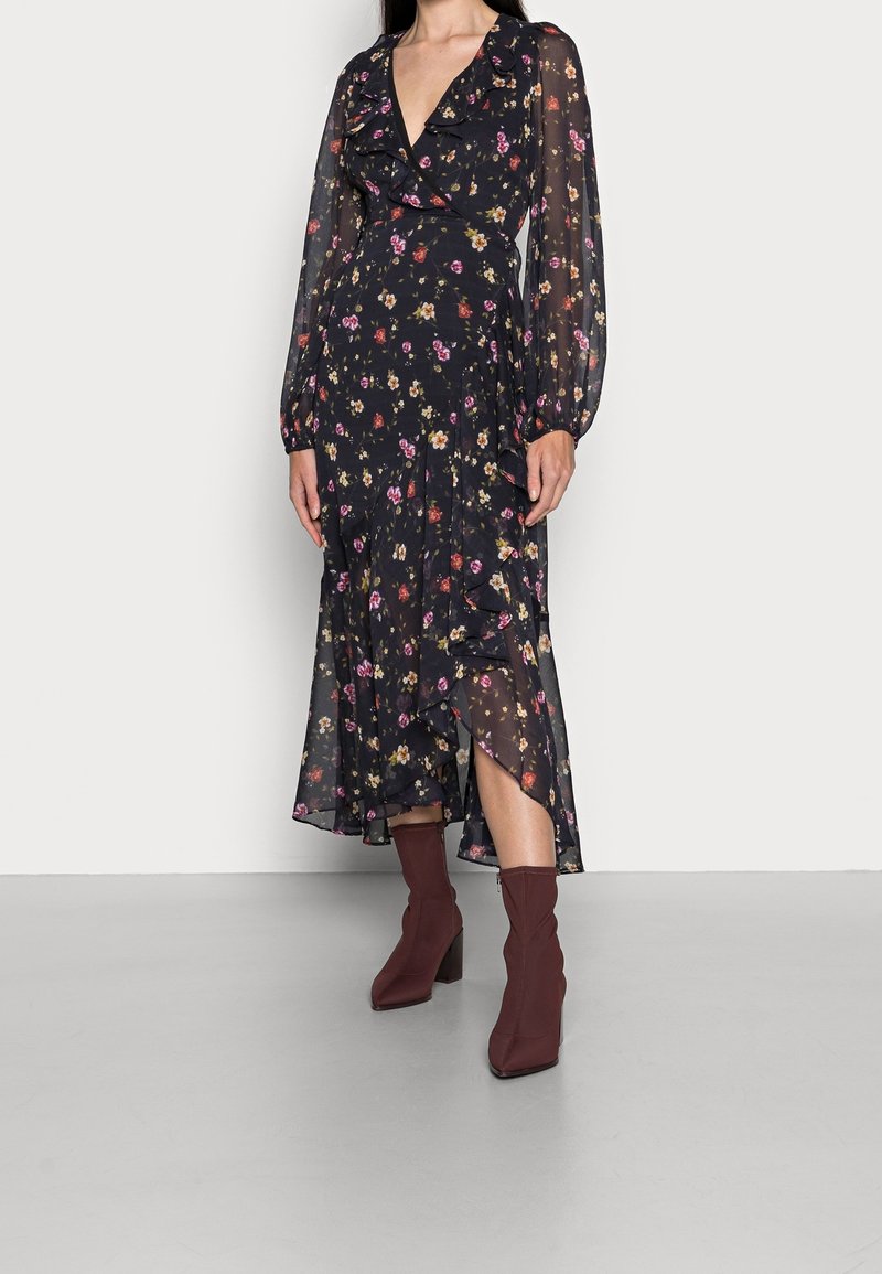 Femme portant une robe portefeuille noire transparente à motifs floraux avec des volants et des manches longues, associée à des bottines rouges foncées.