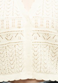 Cardigan blanc en tricot avec des motifs au crochet, boutonné à l'avant, et ourlet festonné, porté avec un jean bleu clair.
