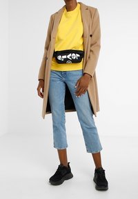 Person som bär en kamelfärgad rock, en gul sweatshirt, avklippta blå jeans, svarta sneakers och en svart midjeväska med vit text.