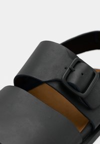 Μαύρο δερμάτινο σανδάλι με φαρδιές λουρίδες και τετράγωνη πόρπη. Διαθέτει μαλακό, καφέ πάτο και υφή στην επιφάνεια.