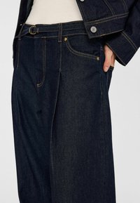 Mörkblå denimbyxor med avslappnad passform, som har hög midja, synliga sömmar och sidofickor. Materialet verkar hållbart och texturerat.