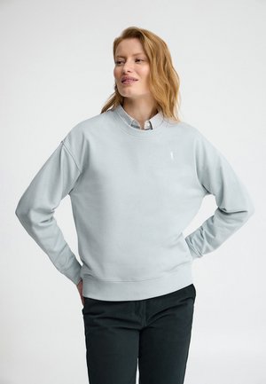 Femme aux cheveux roux clair portant un sweat-shirt gris clair par-dessus une chemise rayée à col et un pantalon noir, les mains sur les hanches, regardant sur le côté.