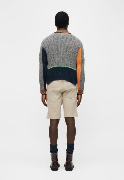Άνδρας που στέκεται με την πλάτη προς το μέρος μας, φορώντας πολύχρωμο πλεκτό πουλόβερ, μπεζ σορτς, σκούρες κάλτσες και καφέ δερμάτινα μποτάκια σε απλό φόντο.