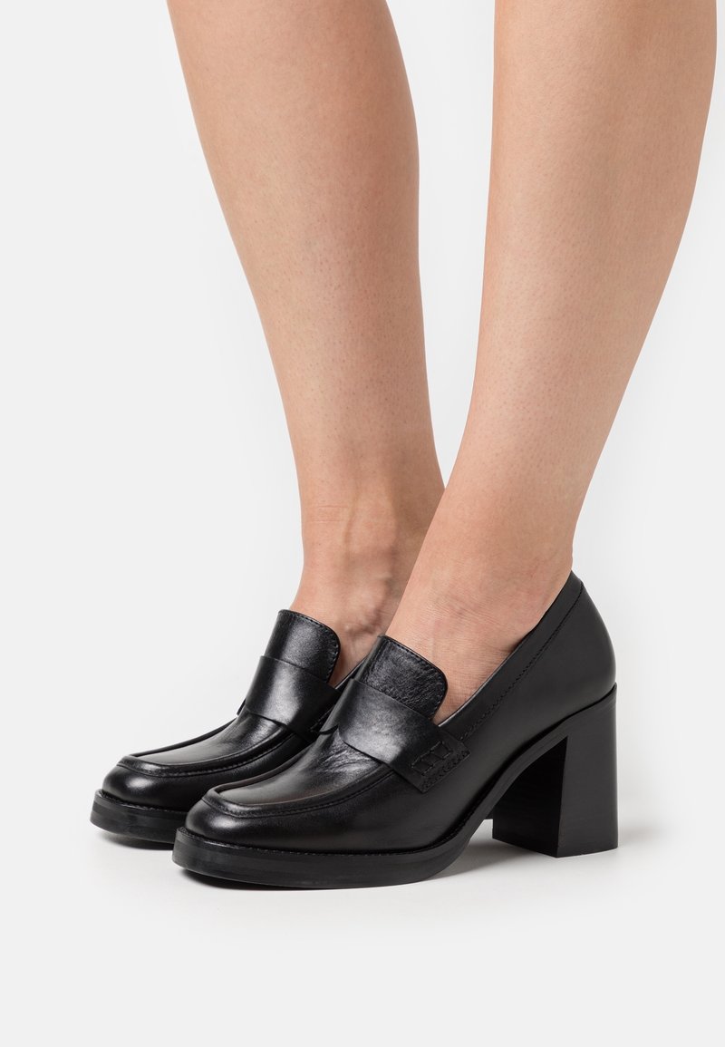 Black leather loafers with a block heel, featuring a smooth finish, rounded toe, and a wide strap across the front.