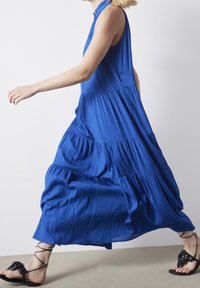 Robe maxi sans manches bleue avec un design à niveaux, une texture plissée et un col haut. Portée avec des sandales noires à lanières ornées d'accents floraux.
