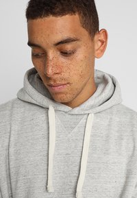 Light gray hoodie made of soft fabric, featuring a front kangaroo pocket, adjustable drawstring, and contrasting cream-colored cords.
