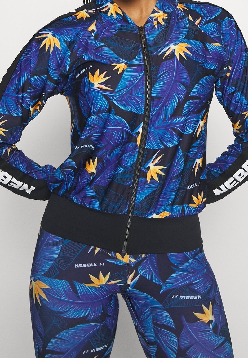 Femme portant une veste zippée et des leggings assortis à imprimé de feuilles tropicales bleues et jaunes avec le logo "NEBBIA" sur les manches et les pantalons.