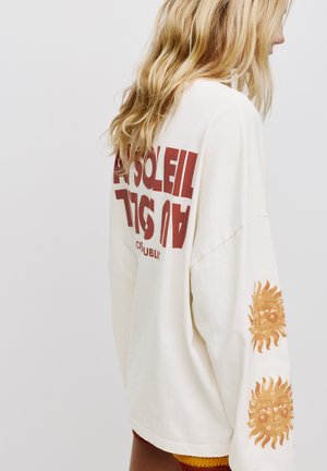 Creme oversized sweatshirt med iøjnefaldende rød "SOLEIL" tekst på ryggen og solmønstre i orange på begge ærmer. Blødt bomuldsstof.