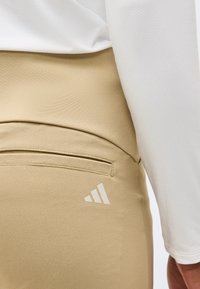 Κοντινό πλάνο μπεζ παντελονιού με πίσω τσέπη και λευκό λογότυπο, φορεμένο με μακρυμάνικη λευκή υφασμάτινη μπλούζα.