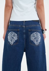 Vaqueros de mezclilla azul oscuro con piernas anchas, que presentan dos diseños de cabezas de león bordados en blanco en los bolsillos traseros. Textura suave con costuras sutiles.